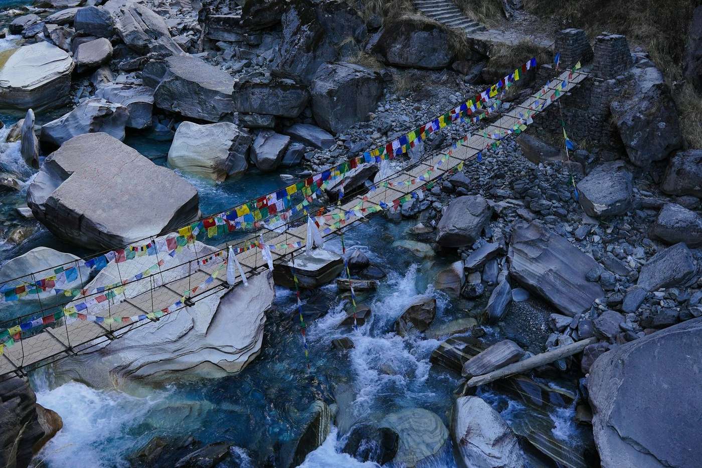 Prayer flag covered bridge over a fast-flowing Himalayan river with large boulders during monsoon