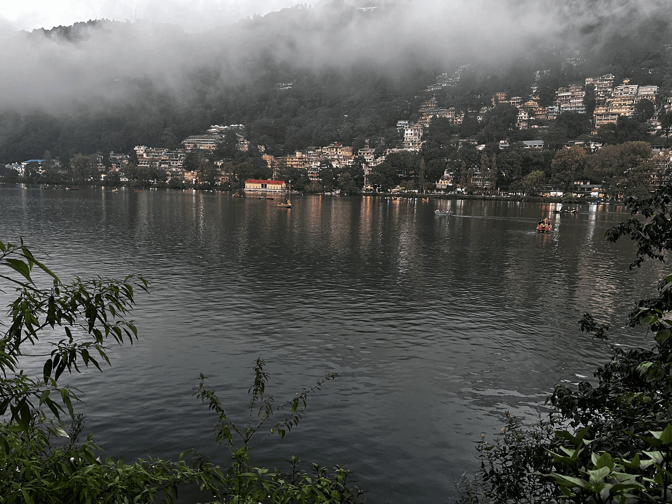 Mist descending over the forested hillside above Naini Lake while boats cross the water